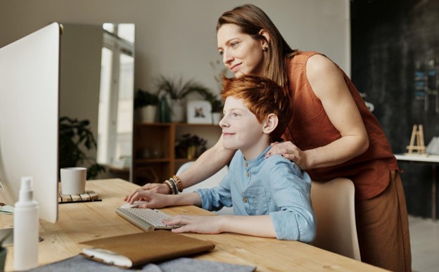 A woman watches her son as he does his homework on a computer.